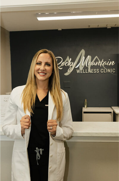 Kylee Johnson is standing in front of the front desk smiling. She's wearing her navy blue scrubs and a white lab coat.