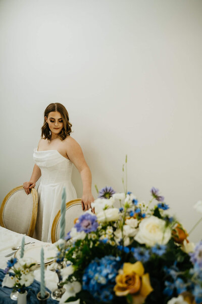A bride stands behind a table with a large floral arrangement, representing wedding management offerings by Birdie June Events and Rentals. 