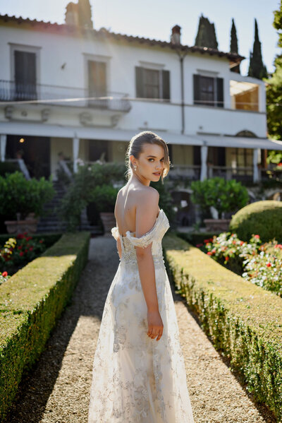 A bride in a lace gown stands on a stone path in a garden with a white villa behind her