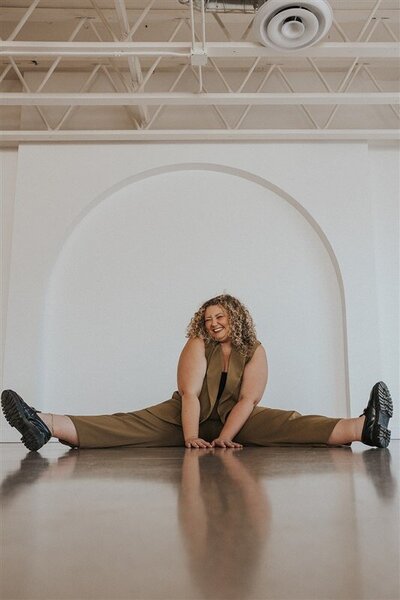 Andrea Cakmar sits in a wide-legged split on the floor, smiling brightly in a studio with a large white arch behind her. She is dressed in a tan suit with a black crop top.