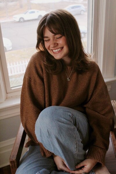 Katie Graper, Showit Web Designer, sitting on a chair smiling with eyes closed while holding her knee to her chest