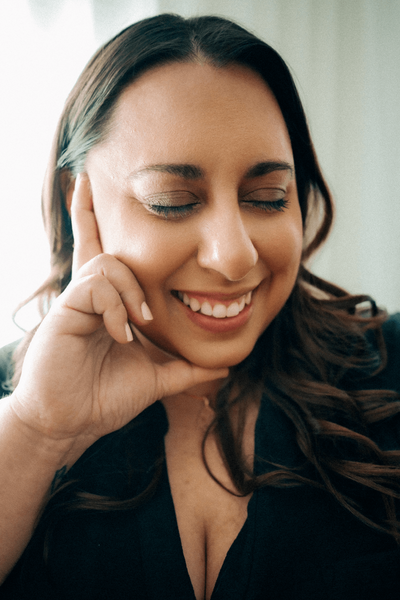 The founder of Nora. Magazine smiling with long brown hair, eyes closed, resting cheek on hand. Wearing a dark top in a softly lit environment, conveying joy and calm.