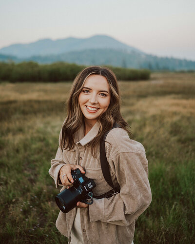 woman standing outside smiling and holding camera with hills in the background