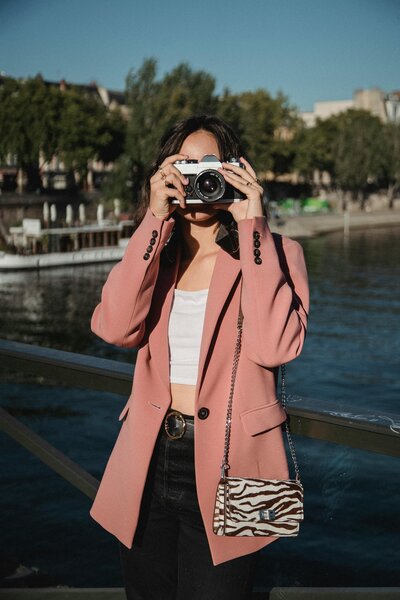 A woman in a pink coat holding a camera in an outdoor setting.
