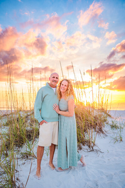 Couple standing in front of a gorgeous golden sunset with a dune and sea grass behind them on bean point, on anna maria island.