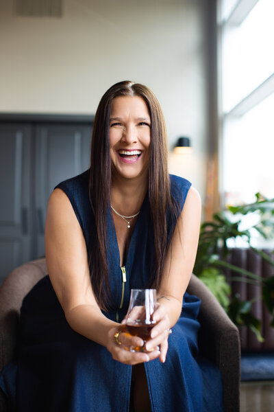 Rawnica Dillingham of New Riff Distillery laughing warmly while seated in a modern Cincinnati studio, holding a glass of whiskey, with natural light streaming through large windows and lush plants in the background — professional personal branding portrait by Janel Lee Photography.