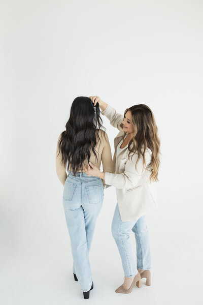 A hairstylist is standing next to her client, showcasing the long dark hair extensions that her stylist is wearing