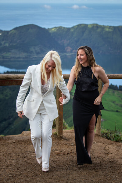 Two women smiling and holding hands during their elopement in the Azores, walking along a scenic overlook with lush green mountains and a lake in the background. One is wearing a white suit, the other a black dress.