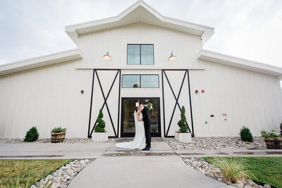 bride and groom kiss in front of white barn at woodlands