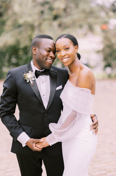 A couple dressed in wedding attire, with the groom wearing a black tuxedo and the bride in an off-shoulder white gown, smile and hold hands outdoors, perfectly epitomizing the vision of partial wedding planning by a seasoned wedding planner Banff.