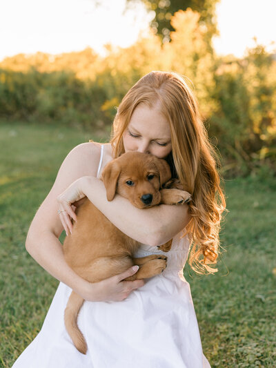 A young woman with red hair embraces a golden retriever puppy in a sunlit field, radiating warmth and happiness. Captured at golden hour by Katie Crane Photography in Toronto, Ontario.