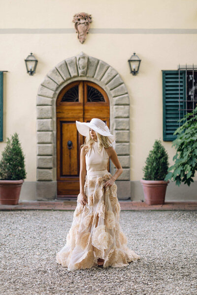 A woman posing in a long floral gown with a hat in front of and arched door with stone.