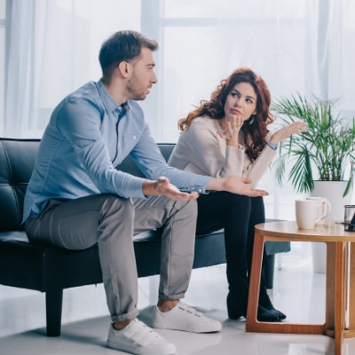 Couple sitting on a sofa in an office both gesture with their hands as they look at each other accusingly. This could represent a disagreement between partners. We offer support with marriage counseling in Miami, FL, couples therapy, and other services.