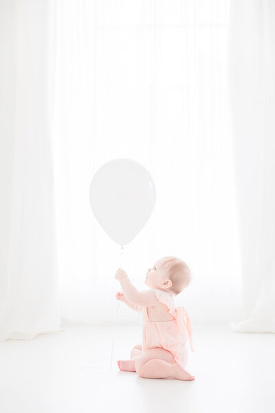 One year old baby girl wearing a pink romper holding balloon in an all white studio