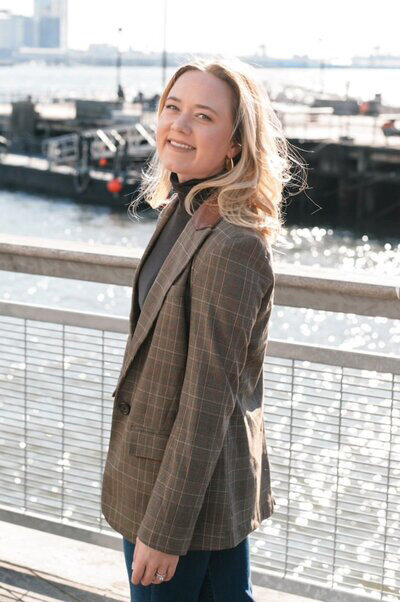 A woman in a blazer and jeans stands confidently on a bridge, representing Olivia Verhulst, Media Consultant.