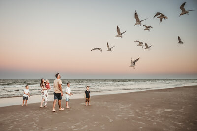 A family with young children are playing on the beach in Corpus Christi during sunset as they watch a flock of seagulls fly overhead.