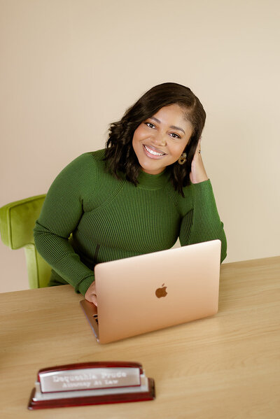 A smiling attorney in a green sweater sitting behind a laptop, with a nameplate reading 'Dequeshia Prude, Attorney At Law' on the desk.