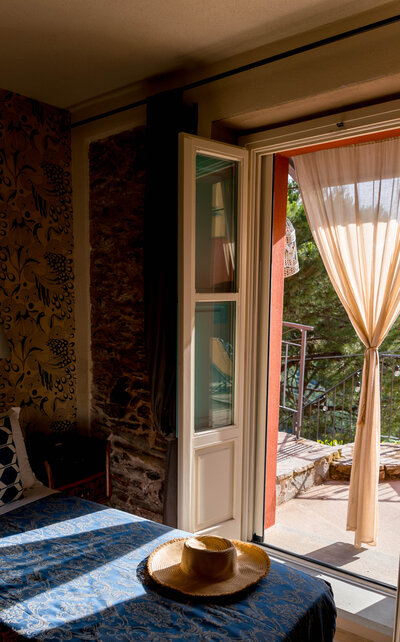 Hotel room with balcony doors open, blue bedding, and a straw sun hat sitting on the bed.