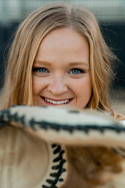 Image of softball catcher holding her mit up and smiling at the camera from behind it.