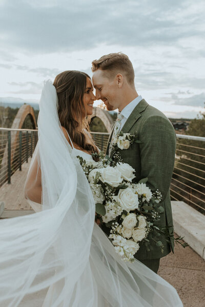 Bride and groom smiling while holding a bouquet, with veil flowing in the wind