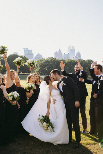 Couple kissing on their wedding day while the wedding party cheers behind and around them.