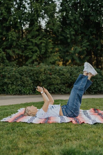 Women laying down on a blanket in the grass  while on her phone with her legs in the air