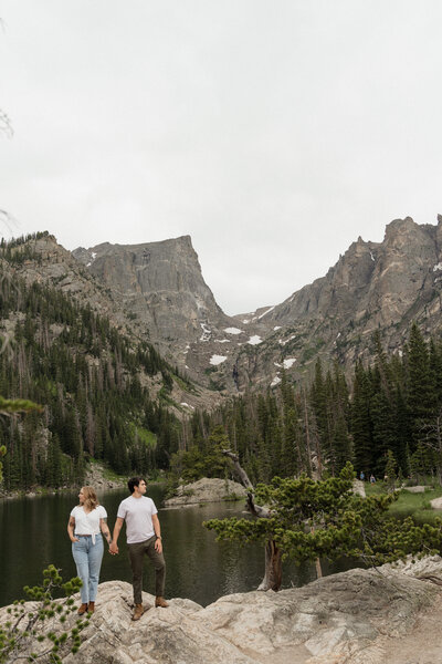 man and woman standing on a cliff over a lake with mountains in the background