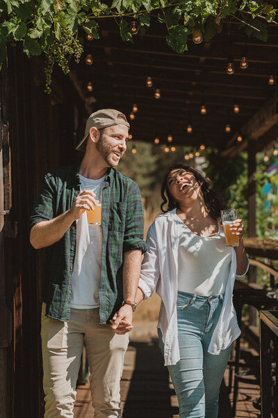 A happy couple strolls under a vine-covered pergola with hanging string lights, each holding a glass of beer. The man, wearing a cap, white t-shirt, and green flannel, smiles as he looks at the woman, who is laughing and wearing a white blouse, denim jeans, and has her hair down. They are holding hands, enjoying a sunny day in a setting that suggests a relaxed outdoor dining or winery environment.