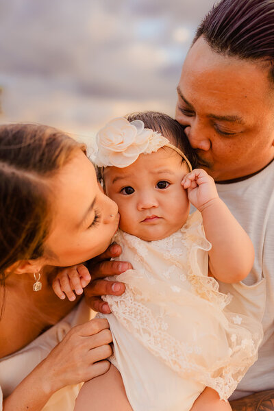 baby looks at camera while mom and dad kiss her head for their beach family photoshoot