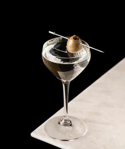 A cocktail glass with clear drink with olive on top with toothpick