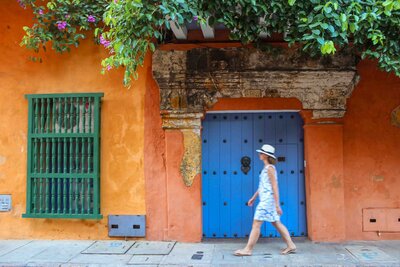 Luxury travel advisor Stephanie Dosch walking past a colonial house with orange walls, blue wooden door, and green window in Cartagena, Colombia ©Stephanie Dosch | theViatrix Luxury Travel