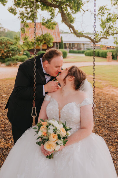 Two people lean in for a kiss on their wedding day, one of them sitting on a swing