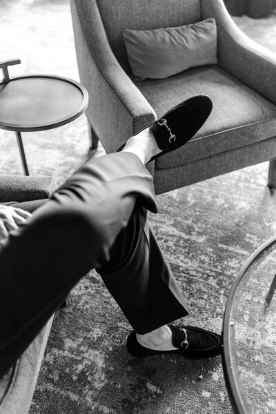 Black and white image of a man seated in a chair with his legs crossed, featuring suit pants and loafers