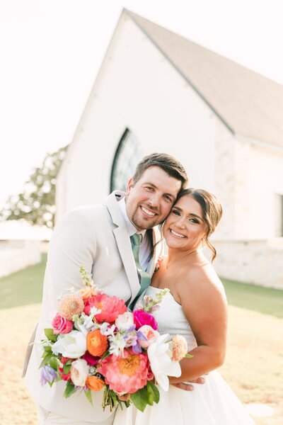 Bride and groom holding colorful floral bouquet outside white wedding venue
