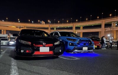 A row of luxury supercars including a Maserati and Ferrari parked under a vibrant Tokyo sky at the iconic Tatsumi Parking Area meet-up spot.