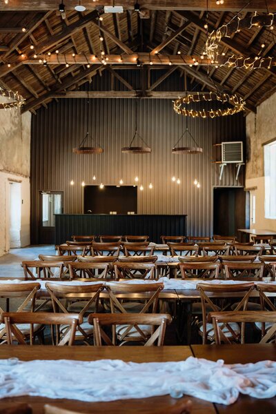 An old barn interior at Kinclune House is photographed. The space is newly renovated and is set up to host 120 wedding guests with rustic wooden chairs and tables.