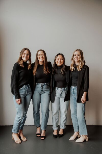 Four women standing smiling with black sweaters and jeans
