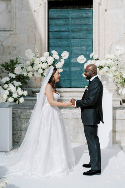 Bride and groom holding hands and smiling during wedding ceremony in front of blue doors and white florals