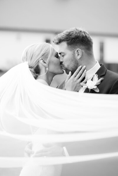 A black and white photo of a groom kissing his bride under a long veil