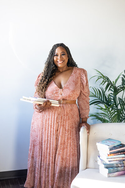 romance author brittainy cherry holding three white books while smiling in a pink paisley dress