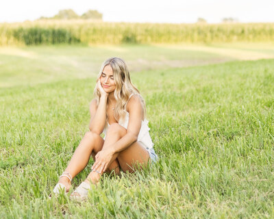 Blond hair female sitting in grass, hand on her chin looking down, corn field in the background