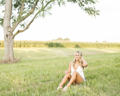 high school senior female, with long blonde hair, sitting in the grass with a tree and cornfield behind her, looking at the camera smiling, wearing blue jean shorts, sandals, and a white spaghetti strapped shirt