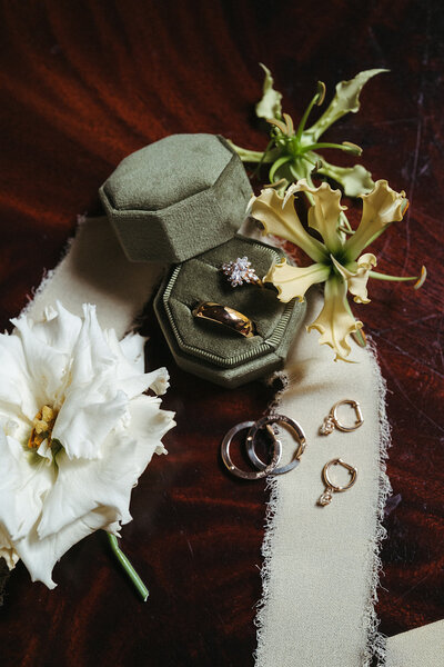Flatlay featured wedding rings in velvet olive box alons with green and white flowers on a rich, dark wood surface.