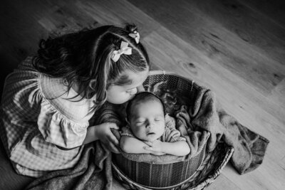 A little girl kissing his newborn brother that is peacefully sleeping in a cozy basket, surrounded by soft blankets.