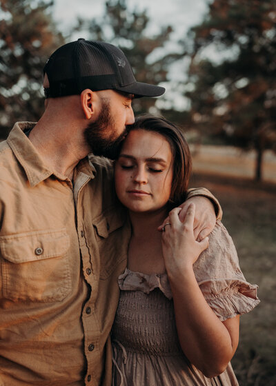 The setting is sunset and a husband has his arm around his wife's shoulder while wearing a baseball cap and kissing her temple.