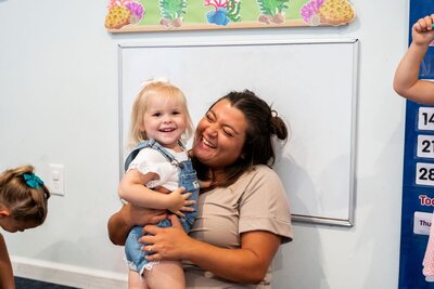Ms. Mia holding a young toddler student as they smile and laugh at The Nest, a natural learning center in Surf City, NC