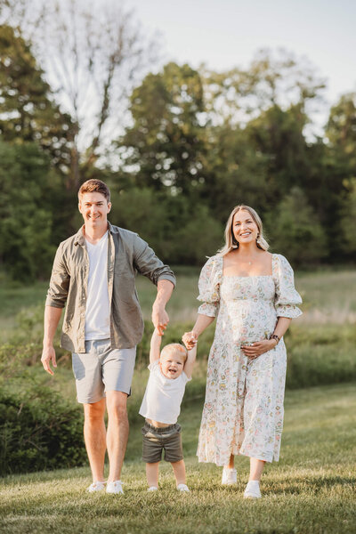 A smiling family of three enjoys a walk in a grassy field during a newborn photography session. The father holds the hand of their excited toddler, who is joyfully lifting his feet off the ground, while the mother, visibly pregnant, walks beside them, holding her own belly and smiling. The setting features lush greenery and a soft, warm light.