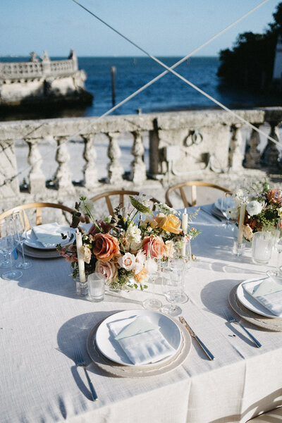 Tablescape from a waterfront reception dinner with grey placesettings and a peach floral centerpiece in the middle