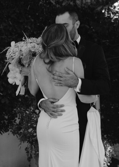 Bride with open back dress holding bouquet and kissing husband in front of bushes