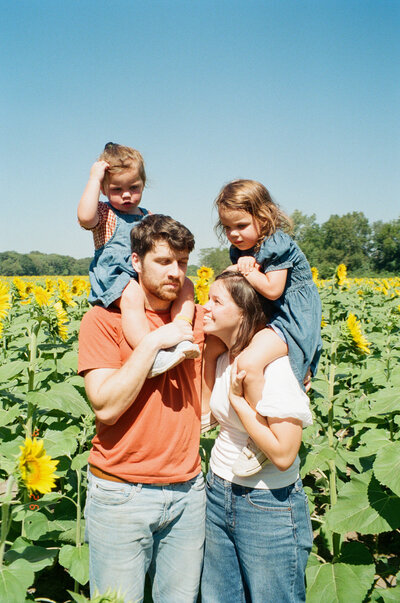 family poses for family photo in sunflower field. kids on parents shoulders